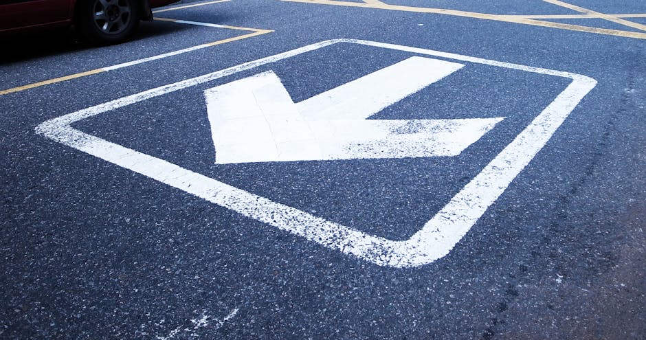 A white-painted downward-pointing arrow within a square on an asphalt surface indicating a designated parking bay for moving vehicles, with adjacent parking lines and the rear wheel of a vehicle visible in the background. The scene is outdoors under natural daylight, likely on a street or designated loading area, relevant to house removals, packing, and furniture transport services provided by Movers Merton for booking a removal parking bay with Merton Council, Merton. This area is used during home relocation or moving logistics to facilitate efficient loading and unloading of household furniture and boxes, supporting the process of packing and moving goods from a property to a moving truck or van.