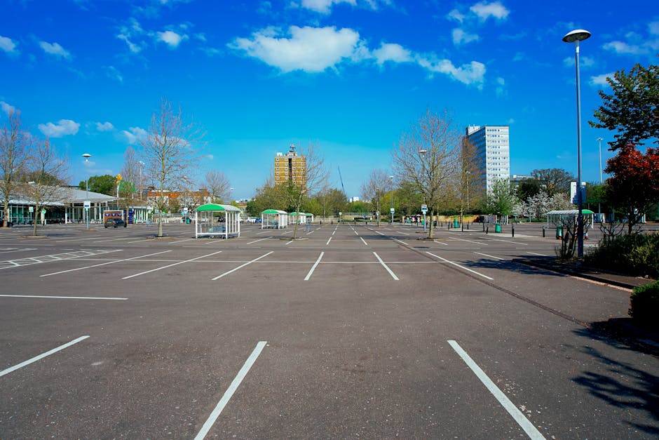 A large, empty parking lot with clearly marked white parking spaces on asphalt surface, situated outdoors during daytime. The lot is surrounded by leafless and blooming trees, some with reddish foliage, and is illuminated by tall street lamps. In the background, there are multiple multi-storey office or residential buildings and a low, modern structure with a glass facade. The sky is bright blue with scattered white clouds. Near the lot’s entrance, there are green and white sheltered bins, possibly for waste or recycling, and a few vehicles parked in the distance. The scene suggests an area used for parking during house removals or furniture transport, with the spacious environment suitable for loading and unloading furniture, boxes, and appliances as part of a home relocation process. Movements such as loading furniture onto vans or trucks could be taking place nearby, supported by the presence of a professional removals company like Movers Merton.