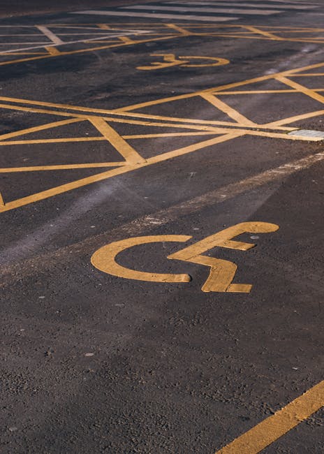 A white-painted downward-pointing arrow within a square on an asphalt surface indicating a designated parking bay for moving vehicles, with adjacent parking lines and the rear wheel of a vehicle visible in the background. The scene is outdoors under natural daylight, likely on a street or designated loading area, relevant to house removals, packing, and furniture transport services provided by Movers Merton for booking a removal parking bay with Merton Council, Merton. This area is used during home relocation or moving logistics to facilitate efficient loading and unloading of household furniture and boxes, supporting the process of packing and moving goods from a property to a moving truck or van.
