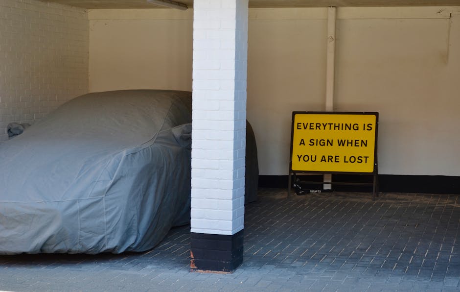 Inside a parking area or garage, a large vehicle or moving van is partially visible with its side door open, showing a load of household furniture and boxes prepared for a home relocation. A sofa or armchair, covered with a grey protective blanket, is positioned on the floor near a white brick pillar. To the right, a bright yellow sign with black text reading 'EVERYTHING IS A SIGN WHEN YOU ARE LOST' is propped upright against the wall. The floor is tiled with dark bricks, and the environment appears organized for a furniture transport or packing and moving process. The setting suggests an early stage of a house removal, with packing materials and furniture ready for loading into the van, facilitated by professional movers such as Movers Merton.
