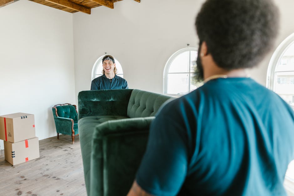 Inside a residential property during a house move, a professional removal team from Movers Merton is assisting with packing and furniture transportation. The image shows a man with curly hair and a beard, wearing a navy blue shirt, standing near the doorway and holding a large, green upholstered sofa which appears to be in the process of being moved. Another team member, a woman with shoulder-length hair and a headband, is visible inside the room, smiling and communicating with the man. The room features light-colored walls, two arched windows allowing natural light to illuminate the space, and a wooden ceiling beam. In the background, there are a few cardboard boxes with red tape, a small vintage-style green armchair, and a spacious, uncluttered environment ready for the packing and loading process. The scene captures the careful handling of furniture and the organized approach typical of home relocation services provided by Movers Merton.
