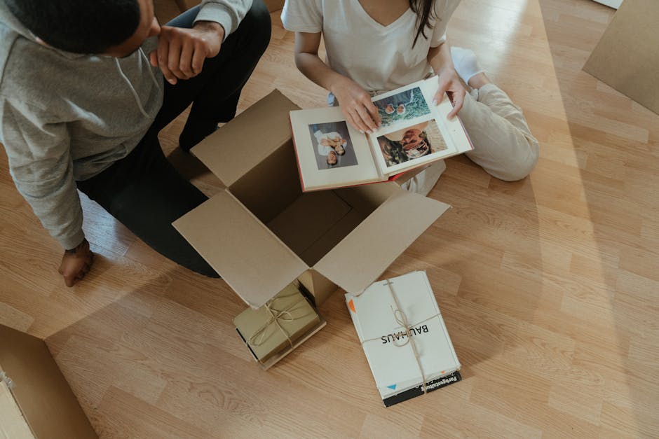 Two individuals are engaged in packing or unpacking household items inside a room with wooden flooring. One person, seated cross-legged, is holding an open photo album or scrapbook featuring several photographs. The other person, kneeling, is partially visible and appears to be assisting or observing. Around them are cardboard boxes, some sealed with packing tape and others open, with one box from [COMPANY_NAME] labeled for moving purposes. A clear plastic bag or wrapping material is visible, likely for protecting fragile items during the home relocation process. The scene suggests an unpacking phase in a furniture transport or packing and moving service, with focus on carefully handling personal belongings as part of a house or apartment move, handled by [COMPANY_NAME]. The environment is well-lit, with the interaction taking place on a wooden floor, emphasizing the preparation and organization involved in successful house removals and logistics.