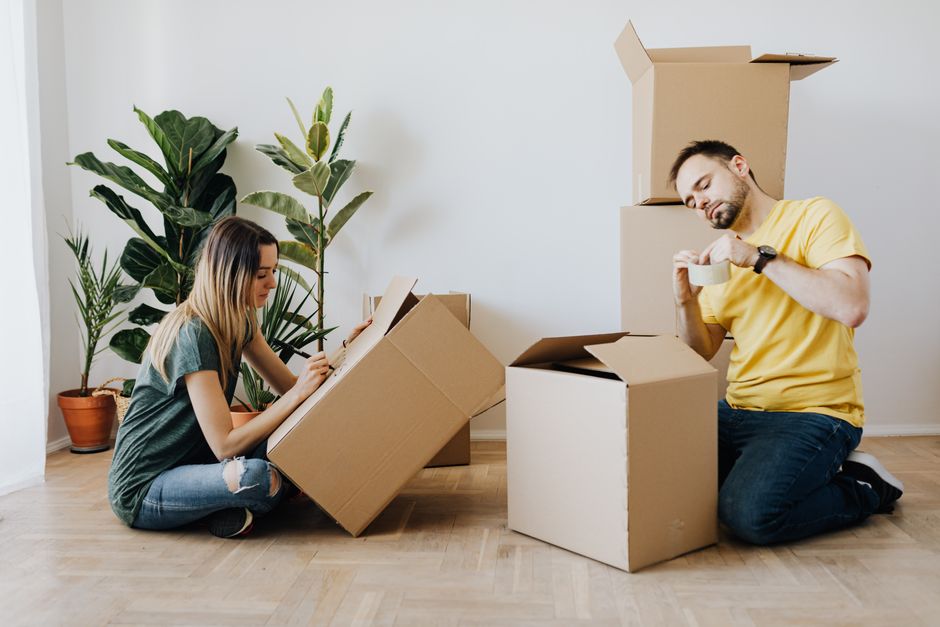 Inside a residential property during a house move, a professional removal team from Movers Merton is assisting with packing and furniture transportation. The image shows a man with curly hair and a beard, wearing a navy blue shirt, standing near the doorway and holding a large, green upholstered sofa which appears to be in the process of being moved. Another team member, a woman with shoulder-length hair and a headband, is visible inside the room, smiling and communicating with the man. The room features light-colored walls, two arched windows allowing natural light to illuminate the space, and a wooden ceiling beam. In the background, there are a few cardboard boxes with red tape, a small vintage-style green armchair, and a spacious, uncluttered environment ready for the packing and loading process. The scene captures the careful handling of furniture and the organized approach typical of home relocation services provided by Movers Merton.