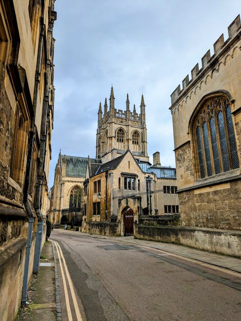 The image depicts a narrow street in an historic city area, with stone and brick buildings lining both sides. On the right, there is a large Gothic-style church with prominent arched windows and ornate stonework, featuring a tall central tower with decorative spires. The buildings on either side have detailed facades, some with large stained-glass windows and traditional wooden window frames. The street is paved with asphalt and has a double yellow line along the curb, indicating parking restrictions. In the distance, the church's tower rises prominently against a partly cloudy sky. This scene illustrates a typical historic city centre setting, potentially relevant to house removals or moving logistics in Merton, where careful planning is required when navigating traditional urban streets with residential and ecclesiastical architecture, as managed by services such as Movers Merton.
