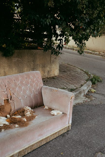 A worn-out, pink upholstered sofa with a tufted backrest and armrest, positioned outside on a paved sidewalk alongside a low concrete wall and a curb. The sofa exhibits significant damage, including torn fabric on the armrest and backrest, with visible stuffing and foam cushions protruding through the tears. Surrounding the sofa, there are small scattered pieces of debris and possible stuffing material. In the background, there is greenery from a tree and bushes, with the sidewalk leading to a street. This scene suggests the sofa has been discarded, possibly as part of home removal or furniture disposal, and may be in preparation for collection by a professional removals service like Movers Merton, specialising in furniture transport and house removals. The lighting indicates daytime, with natural light illuminating the area.