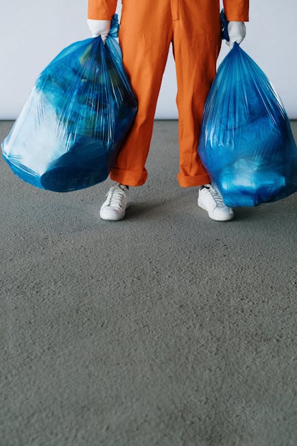 The image shows a person dressed in orange work trousers and white sneakers, holding two large blue plastic bags filled with various items, likely related to house removals or waste disposal. The individual is standing on a grey carpeted indoor floor near an open doorway that leads to a brightly lit room with white walls. In the background, there are several cardboard boxes and a piece of furniture partially visible, suggesting packing or clearance activities associated with home relocation. The scene emphasizes the process of loading and transporting items, with the person engaged in carrying the bags, which are sealed with twist ties or drawstrings. The setting appears to be inside a residential property, preparing for a furniture transport or moving process, consistent with professional services offered by Movers Merton for local removal and disposal solutions in Mitcham and Merton.