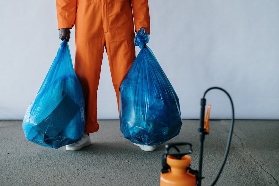 The image shows a person dressed in orange work trousers and white sneakers, holding two large blue plastic bags filled with various items, likely related to house removals or waste disposal. The individual is standing on a grey carpeted indoor floor near an open doorway that leads to a brightly lit room with white walls. In the background, there are several cardboard boxes and a piece of furniture partially visible, suggesting packing or clearance activities associated with home relocation. The scene emphasizes the process of loading and transporting items, with the person engaged in carrying the bags, which are sealed with twist ties or drawstrings. The setting appears to be inside a residential property, preparing for a furniture transport or moving process, consistent with professional services offered by Movers Merton for local removal and disposal solutions in Mitcham and Merton.