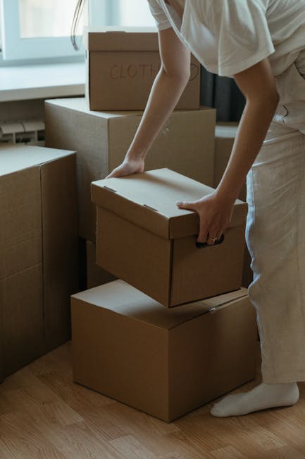 A person wearing light-coloured clothing and white socks is seen in an indoor space, carefully stacking or organizing cardboard boxes, including a small box labeled 'Clothes', on a wooden floor near a window. Several other cardboard boxes of varying sizes are arranged nearby, some stacked, suggesting the process of home packing or moving. The scene takes place in a well-lit room with natural light coming through the window, and the individual appears to be involved in household packing as part of a home relocation carried out by Movers Merton, a professional removals service specializing in furniture transport and packing and moving logistics.