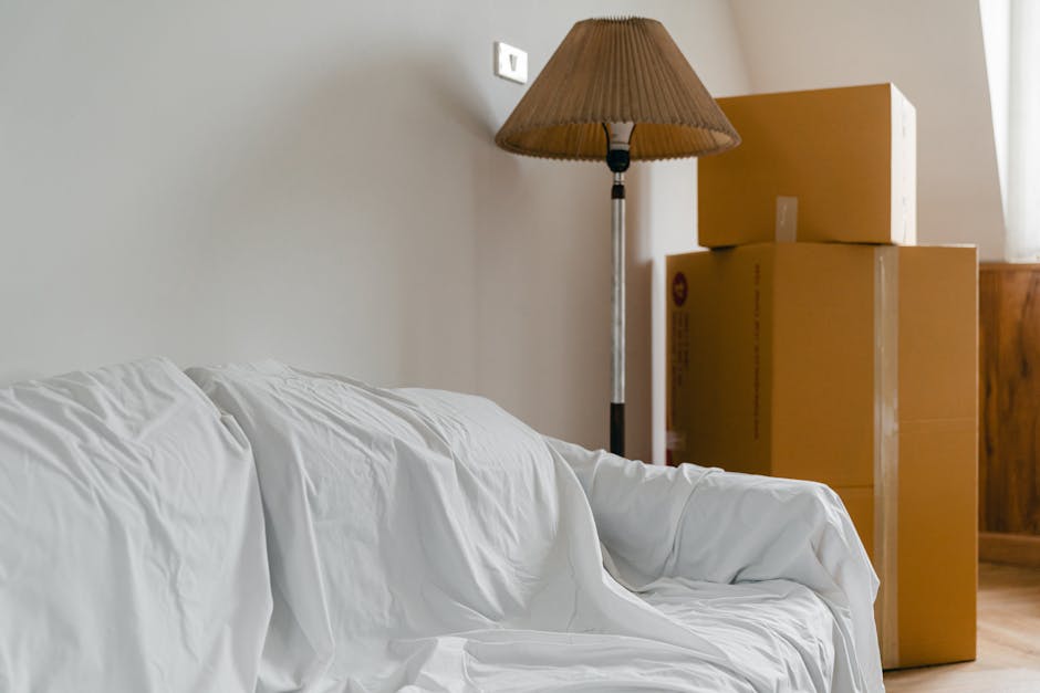 Interior scene showing a white fabric-covered sofa positioned in a room with plain light-colored walls. Behind the sofa, there is a tall floor lamp with a brown pleated lampshade and a metallic stand placed next to a stack of three large cardboard boxes, likely packed for a house move. The boxes are sealed with clear packing tape and vary in size, with two smaller boxes stacked on top of a larger one. The scene appears to be in a home environment, and the boxed items are probably prepared for furniture transport and home relocation services. The natural lighting illuminates the space, highlighting the materials and objects associated with packing and moving processes, with the presence of storage and packing materials hinting at ongoing or planned household removals handled by moversmerton.co.uk.