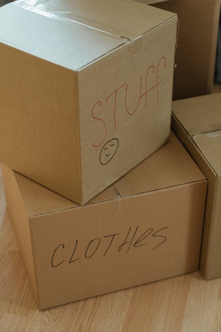A person wearing light-coloured clothing and white socks is seen in an indoor space, carefully stacking or organizing cardboard boxes, including a small box labeled 'Clothes', on a wooden floor near a window. Several other cardboard boxes of varying sizes are arranged nearby, some stacked, suggesting the process of home packing or moving. The scene takes place in a well-lit room with natural light coming through the window, and the individual appears to be involved in household packing as part of a home relocation carried out by Movers Merton, a professional removals service specializing in furniture transport and packing and moving logistics.