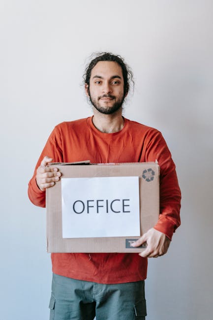 A man with dark, curly hair and a beard is standing indoors against a plain light grey wall. He is wearing a red long-sleeved shirt and grey trousers. He holds a cardboard box with a white label that reads 'OFFICE', supported at chest level with both hands. The box appears to be sealed with packing tape, and the man has a neutral expression on his face. This image is related to office removals and furniture transport, showcasing the process of packing or moving office supplies as part of professional house and office relocation services provided by Movers Merton. The background and lighting are simple and clean, emphasizing the focus on the man and the labeled box, relevant to the themes of packing, loading, and transport during furniture removals or home relocation processes.