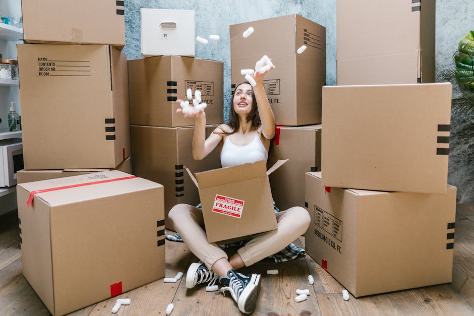 A young woman sitting cross-legged on a wooden floor inside a room, surrounded by numerous cardboard boxes, some sealed with red tape and others open with packing materials visible. She is smiling and tossing white packing peanuts into the air, with some falling around her and into an open box on her lap that has a red 'Fragile' label. Behind her, additional boxes are stacked against a blue textured wall, and a small green plant is partially visible on the right side. The scene depicts a packing or moving process, with various sizes of boxes prepared for a home or office relocation, consistent with furniture transport and packing activities associated with [COMPANY_NAME]'s removals services.