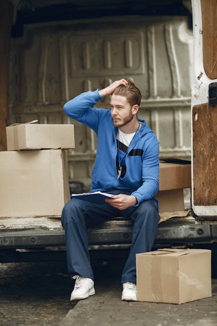 A young man with light brown hair, dressed in a blue hoodie layered over a white t-shirt and dark trousers, is sitting on the edge of a partially open moving van's tailgate in an outdoor setting. He i