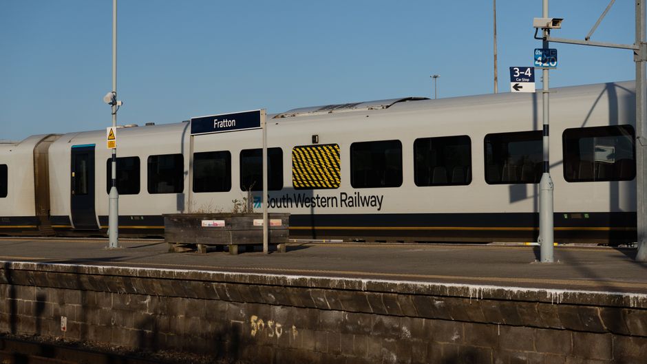 Image of a South Western Railway train stopped at a station platform, with several mobile signs indicating platforms 3-4 and a sign displaying 'Fratton'. The platform surface is made of concrete, with a weathered edge visible in the foreground. Several railway poles with surveillance cameras and warning signs are positioned along the platform, supporting overhead wires. The train has a white and grey exterior with black window frames and a yellow stripe, suggesting it is used for commuter or regional transport. The environment is outdoors under clear blue skies, with natural daylight illuminating the scene. This setting may relate to the logistics of relocating household items, as part of a home or flat clearance service offered by Movers Merton, who specialise in removals and furniture transport near South Wimbledon station, Merton.
