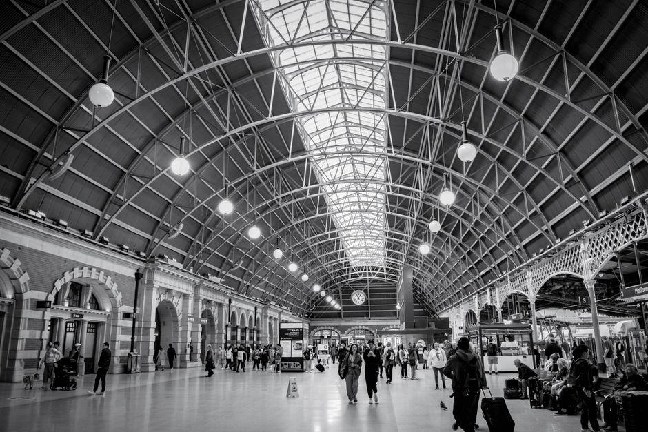 A busy train station interior in black and white, featuring a high arched ceiling with metal framework and large glass panels allowing natural light to filter in. The station is filled with numerous travelers, some walking, others waiting near benches or standing with luggage. Suitcases, backpacks, and shopping bags are visible, and a range of cardboard boxes wrapped in plastic or placed on trolleys are being moved around. Several individuals are seen carrying or pushing boxes and small furniture pieces, indicating a home relocation or furniture transport process. The platform and waiting areas are adjacent to ticketing booths, signage, and a large clock hanging on the wall. This setting is an environment where professional removal services, such as those offered by Movers Merton, might assist with packing, moving, and transport logistics, integrating the logistics of house removals within the station’s busy atmosphere.