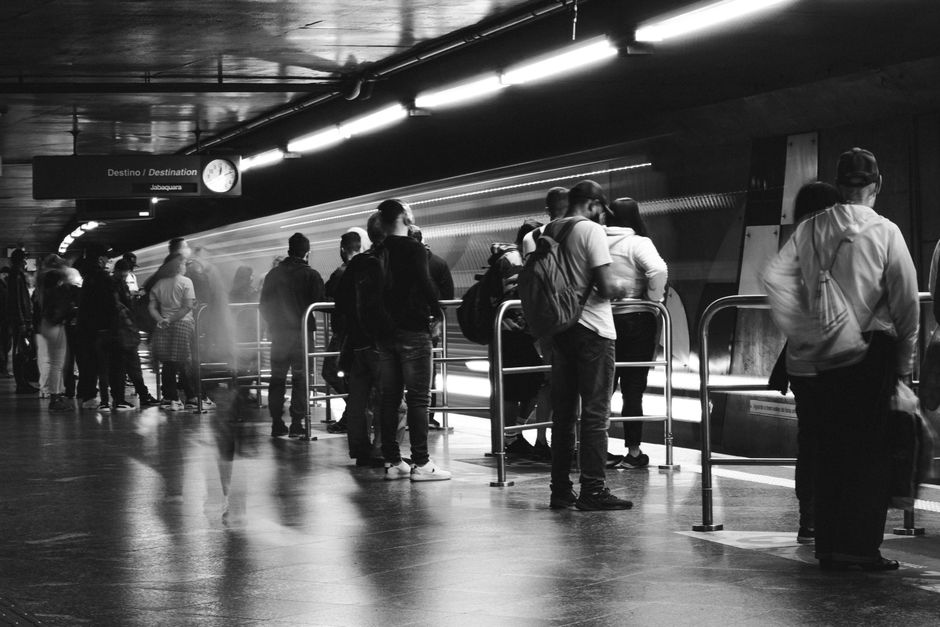 Image of a South Western Railway train stopped at a station platform, with several mobile signs indicating platforms 3-4 and a sign displaying 'Fratton'. The platform surface is made of concrete, with a weathered edge visible in the foreground. Several railway poles with surveillance cameras and warning signs are positioned along the platform, supporting overhead wires. The train has a white and grey exterior with black window frames and a yellow stripe, suggesting it is used for commuter or regional transport. The environment is outdoors under clear blue skies, with natural daylight illuminating the scene. This setting may relate to the logistics of relocating household items, as part of a home or flat clearance service offered by Movers Merton, who specialise in removals and furniture transport near South Wimbledon station, Merton.