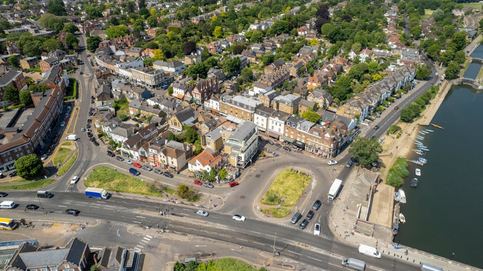 An aerial view of a residential area in Wimbledon, SW19, featuring closely spaced terraced and semi-detached houses with tiled roofs, interspersed with lush green trees and small front gardens. The scene includes a main road with cars and vans, some parked along the sides, and a loading zone where a small white moving van is being loaded or unloaded with furniture and boxes wrapped in plastic and cardboard. Visible within the vicinity are street furniture such as benches and street lamps, adjacent to a river with a paved walkway and small boats moored along the edge. The environment indicates a peaceful day with clear weather, and the image captures elements typical for home relocation and furniture transport processes, reflecting elements associated with professional removals services like those provided by Movers Merton in the context of house removals in Wimbledon.