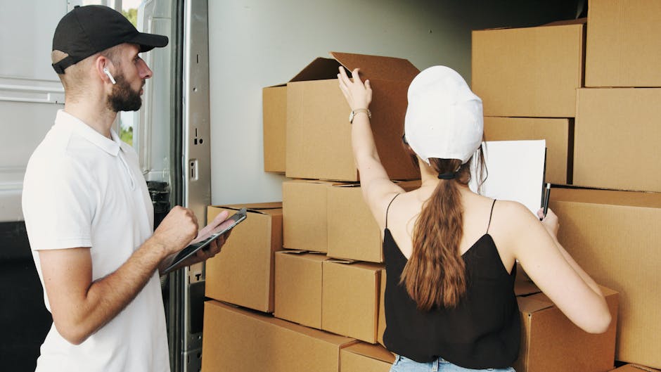 A woman with a ponytail, wearing a white cap, black tank top, and jeans, is standing outdoors near a driveway, holding a tablet and taking notes with a pen. She is surrounded by several cardboard boxe