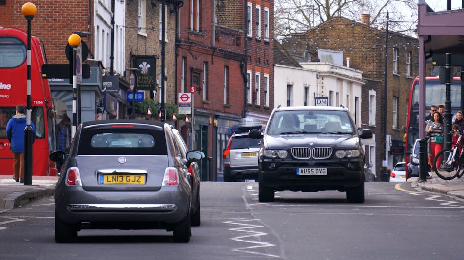A street scene on Wimbledon Common showing parked vehicles, including a grey Fiat 500 with a yellow UK registration plate and a black BMW SUV, with a man wearing a green shirt and dark trousers standing nearby. The street is lined with brick and white terraced houses, some with ground-floor shops, and a red double-decker bus is partially visible on the left. Pedestrians and cyclists are present, with some people waiting at bus stops and others walking along the pavement. Visible elements include traffic signs, bollards, and street furniture. This urban environment illustrates the challenges of home relocation and furniture transport in narrow lanes, where careful loading and moving logistics are required, often assisted by professional removals services like Movers Merton. The scene captures the typical setting for a house removal involving loading furniture and boxes onto vehicles within a busy residential area.