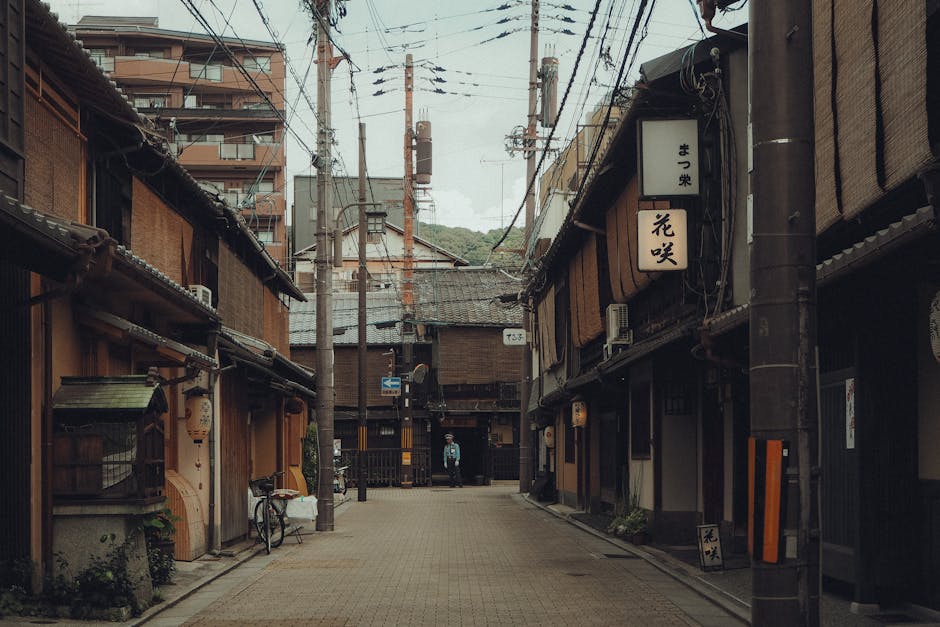 A narrow street scene with traditional Japanese-style wooden buildings lining both sides, featuring sliding doors and paper lanterns, with overhead power lines and utility poles visible above. The street has a paved surface with a bicycle leaning against a building on the left side. In the background, a person wearing a vest stands near an entrance, and there are some potted plants and signage on the buildings. This setting illustrates a compact, historic urban area suitable for house removals and moving logistics services, where careful navigation and furniture transport may be necessary during home relocation. The lighting appears natural with overcast sky conditions, highlighting the detail in the building facades, which are typical of a traditional Japanese neighbourhood, making it relevant for moving services in such environments.