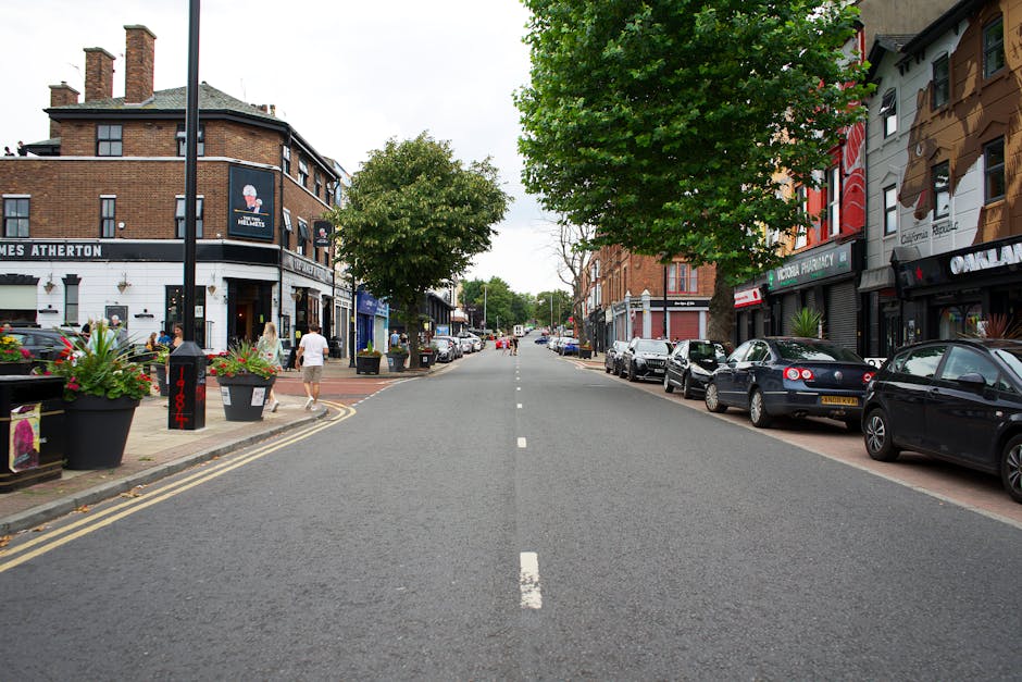 A street scene on Wimbledon Common showing parked vehicles, including a grey Fiat 500 with a yellow UK registration plate and a black BMW SUV, with a man wearing a green shirt and dark trousers standing nearby. The street is lined with brick and white terraced houses, some with ground-floor shops, and a red double-decker bus is partially visible on the left. Pedestrians and cyclists are present, with some people waiting at bus stops and others walking along the pavement. Visible elements include traffic signs, bollards, and street furniture. This urban environment illustrates the challenges of home relocation and furniture transport in narrow lanes, where careful loading and moving logistics are required, often assisted by professional removals services like Movers Merton. The scene captures the typical setting for a house removal involving loading furniture and boxes onto vehicles within a busy residential area.